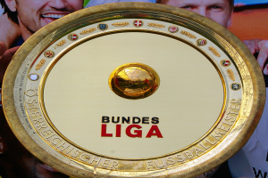 Ein Mann hält einen goldenen Schild mit 'Bundesliga' darauf, im Hintergrund eine Fahne mit drei Personen