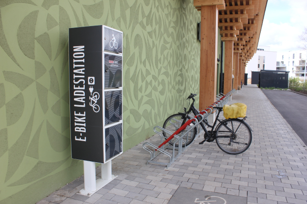 Ein Fahrrad, das vor einem Gebäude mit einem "E-Bike-Ladestation"-Schild geparkt ist, mit einem Rad im Vordergrund und Gebäuden, Bäumen und einem klaren Himmel im Hintergrund.