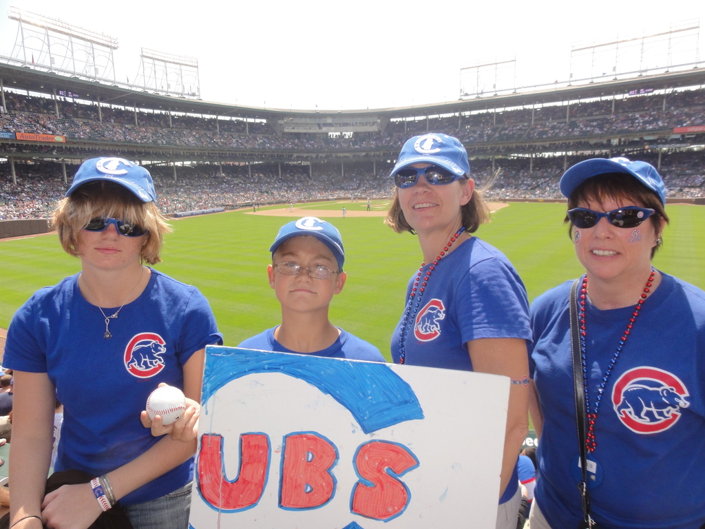 Drei Frauen in Kappen und Brillen, ein Junge in Brille und Kappe mit einem Ball und einer Tafel, stehend in einem Stadion mit Zuschauern und Feld dahinter, unter einem Himmel mit wenigen Lichtern.