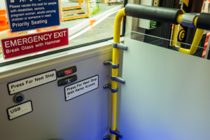 Notausgangsschild an der Seite eines Zuges mit einer Tür, Griff und Glasfenster, das Straße, Pfosten und andere Objekte draußen zeigt.