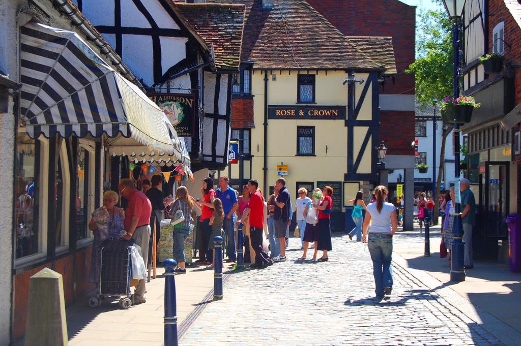 Eine Straße mit Pfählen und Gebäuden auf beiden Seiten, Menschen auf der linken Seite mit einem Handwagen, ein Mann und eine Frau auf der rechten Seite in der Nähe eines lila Müllcontainers, mit Gebäuden, Bäumen und Blumentöpfen im Hintergrund.