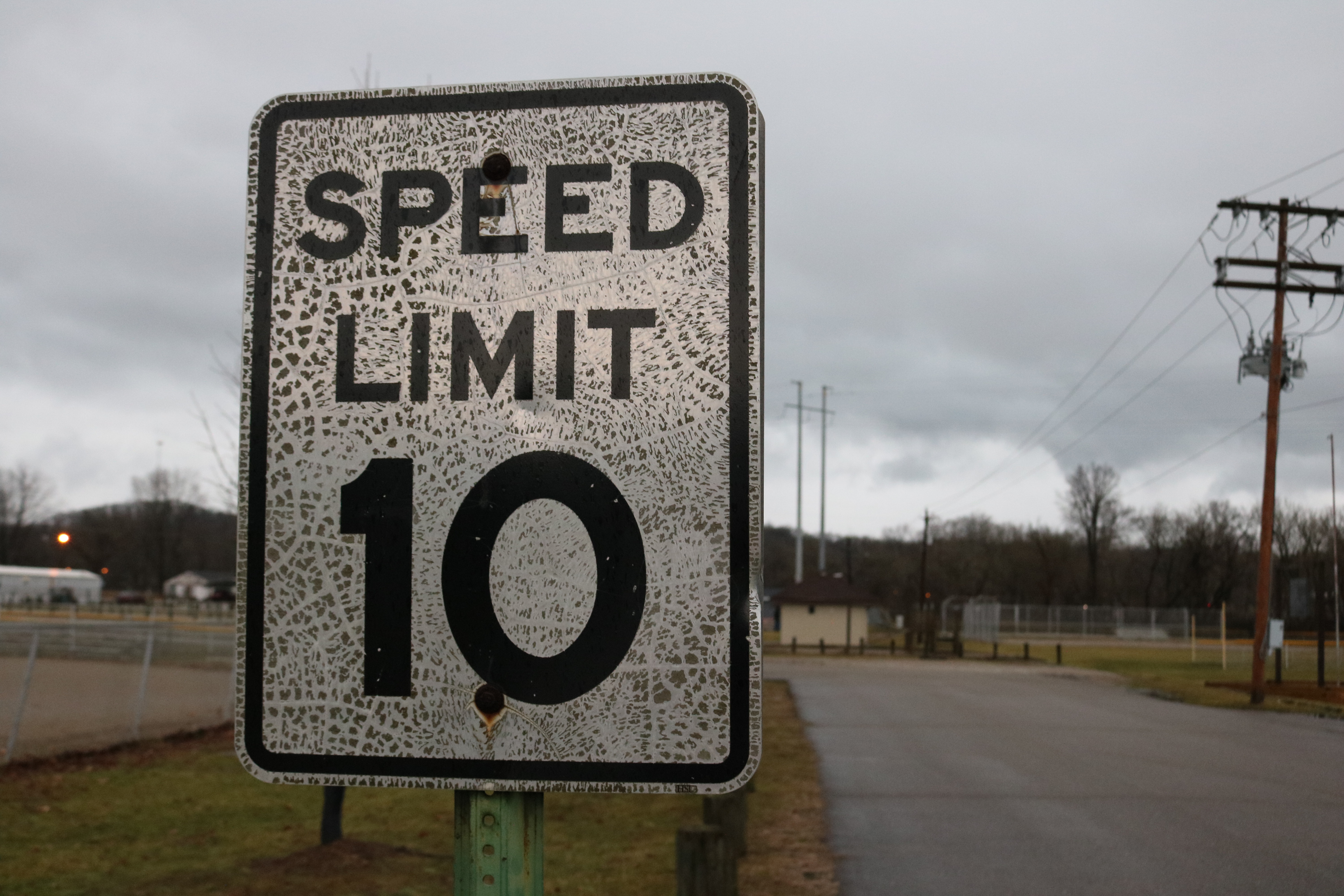 Ein Geschwindigkeitsbegrenzungsschild am Straßenrand, umgeben von Gras, Strommasten mit Drähten, einem Zaun, Häusern, Bäumen und einem bewölkten Himmel im Hintergrund.