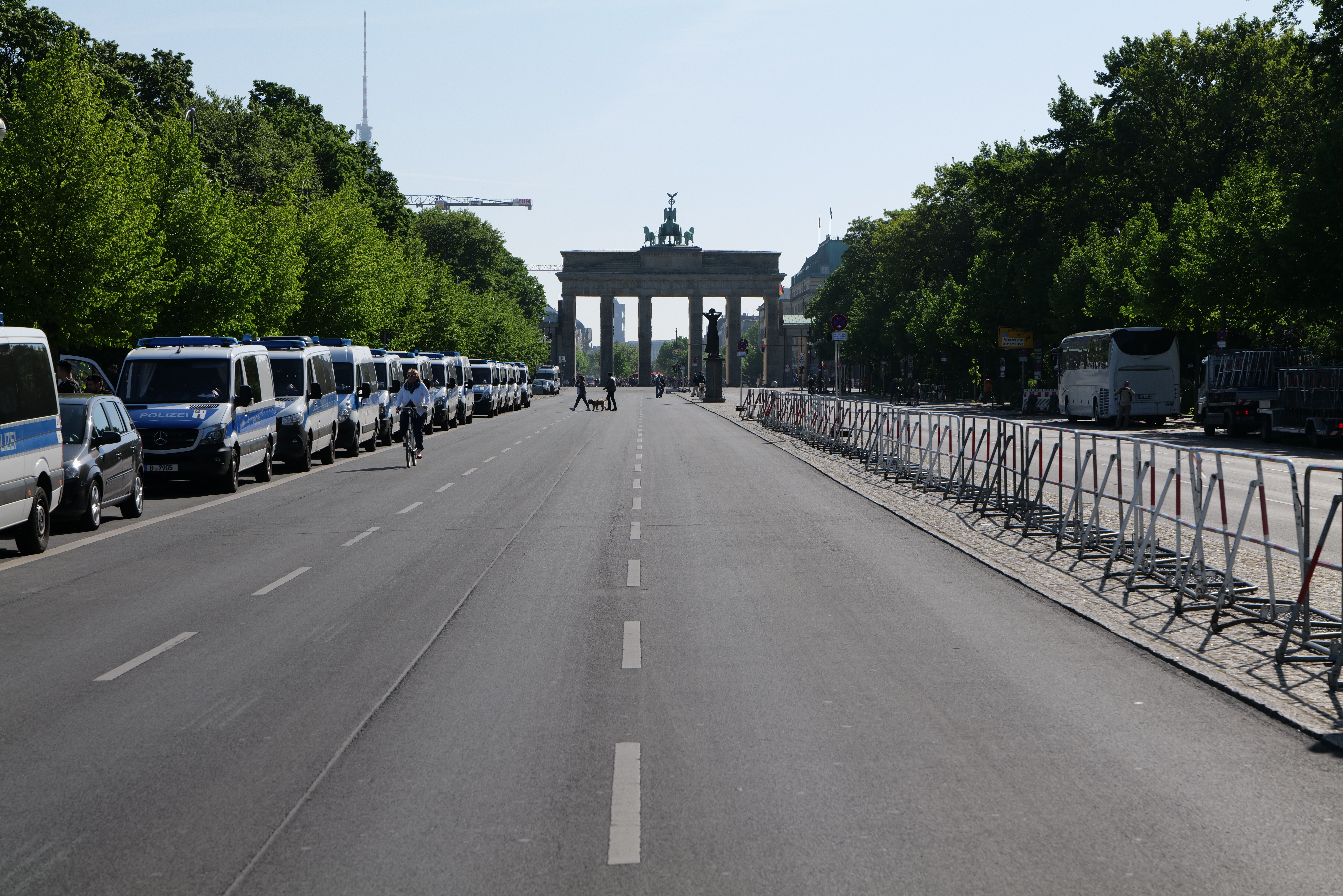 Eine lange Reihe von Polizeiwagen, die auf der Seite einer Stra√üe vor dem Brandenburger Tor in Berlin, Deutschland, geparkt sind, mit Menschen, die Fahrr√§der fahren und auf der Stra√üe stehen, sowie Absperrungen und B√§ume, die die Seiten säumen, und ein Tor mit Statuen im Hintergrund.