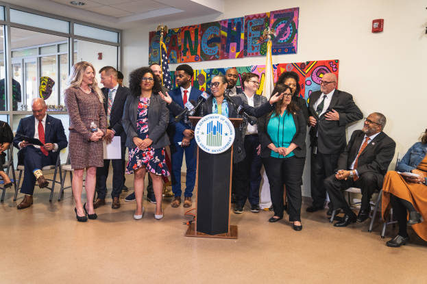 Eine Gruppe von Menschen, die vor einem Podium mit Mikrofon und einer Tafel mit der Aufschrift "Sancho-Stadtrat tritt dem Stadtrat bei" stehen und sitzen; Flaggen, ein Feuerlöscher und Deckenleuchten sind an der Rückwand zu sehen.