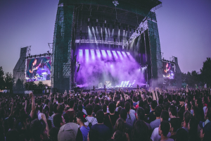 Eine große Menge Menschen auf einem Musikfestival schaut Performern auf einer hell beleuchteten Bühne mit Instrumenten und Bildschirmen zu, vor einem Gebäude, Bäumen und einem klaren blauen Himmel.