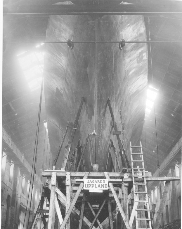 Schwarzes und weißes Foto eines großen Bootes im Bau in einer Werft, das eine Leiter, hölzerne Pfosten, eine Schautafel, Metallrahmen, Deckenleuchten und Fenster zeigt.