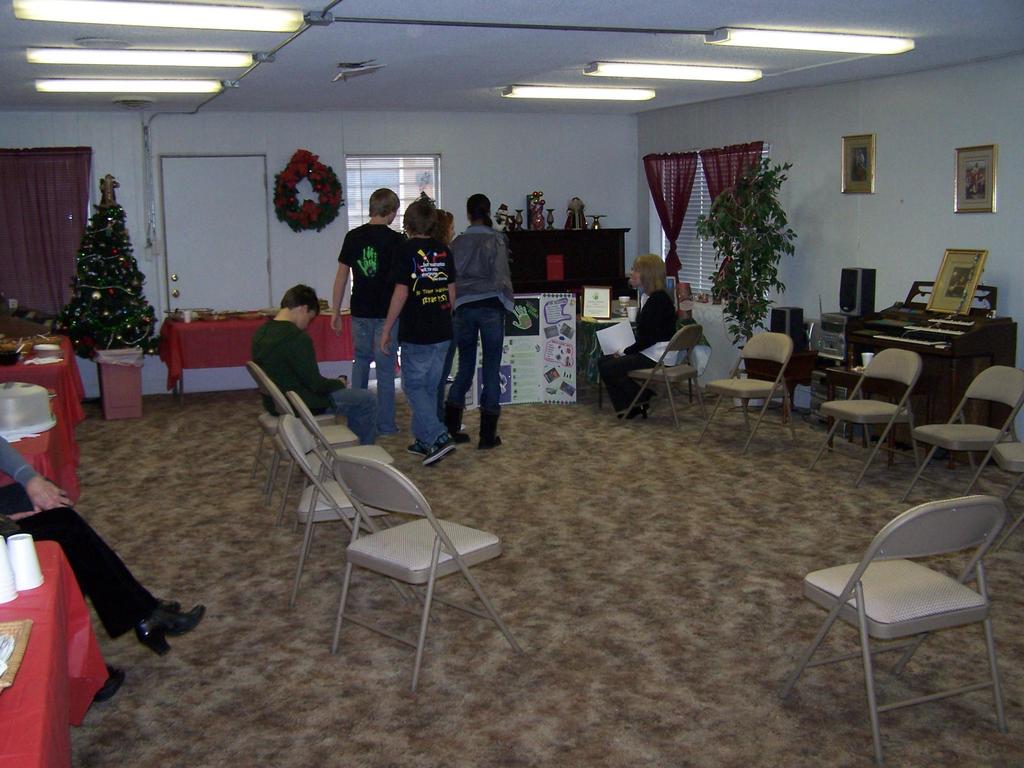 Ein Raum mit mehreren Stühlen, Tischen und Personen, einige stehen, einige sitzen, mit Tassen und Gefäßen auf den Tischen, einem Weihnachtsbaum, einem Piano auf der rechten Seite, Foto Rahmen, Fenstern und Röhrenleuchten an der Decke und einer Wand mit einem Strauß, Vorhang und Tür im Hintergrund.