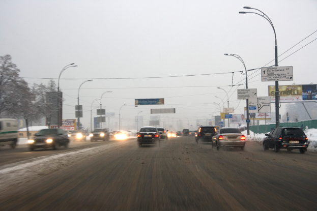 Eine städtische Straße mit viel Verkehr an einem verschneiten Tag, mit Fahrzeugen, schneebedeckter Erde, Laternen, Texttafeln, Bäumen, Gebäuden und einem Himmel im Hintergrund.