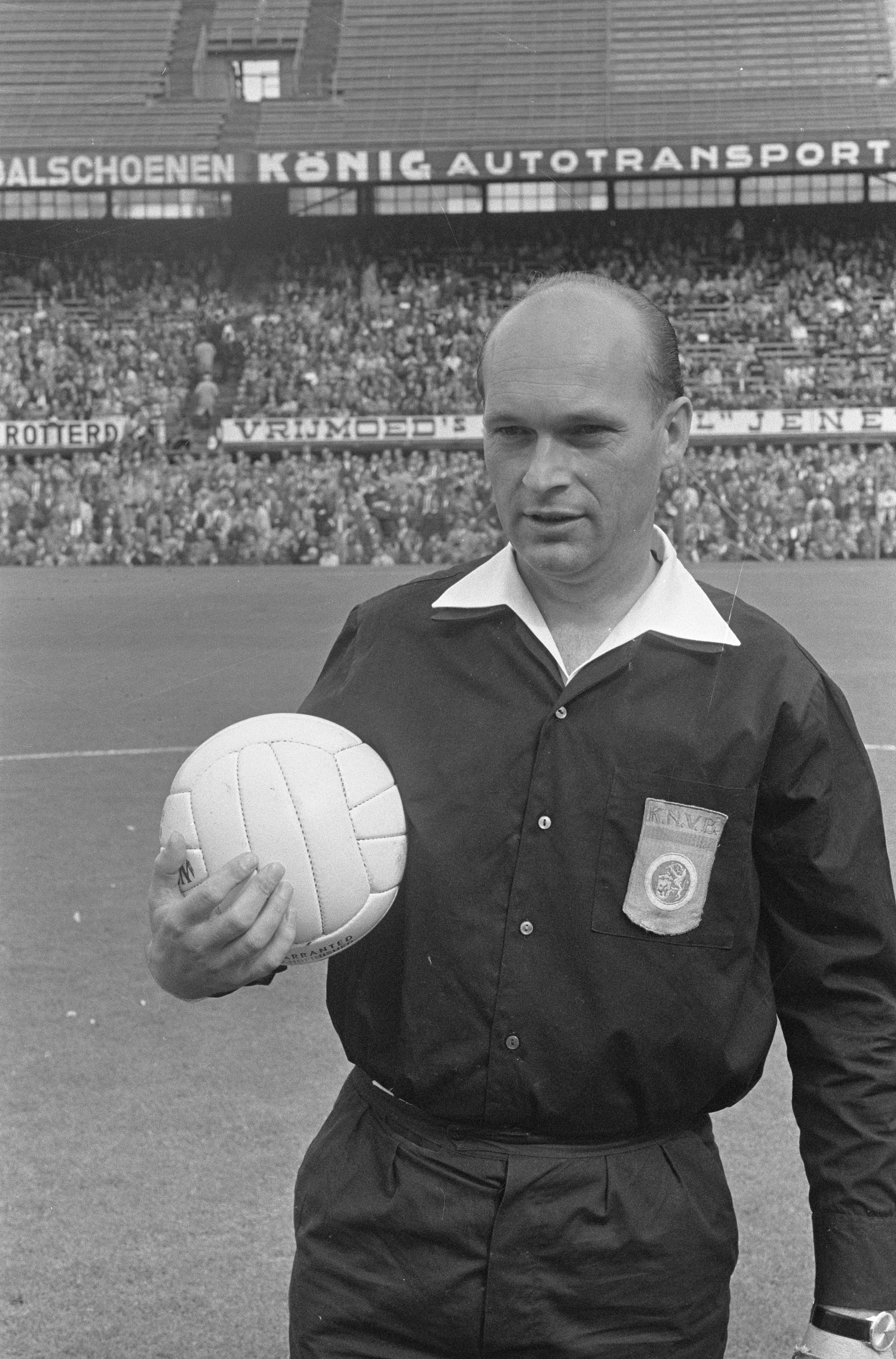 Ein Schwarz-Weiß-Foto eines Schiedsrichters auf einem Fußballfeld, der einen Ball hält, mit Zuschauern und Schildern im Hintergrund.
