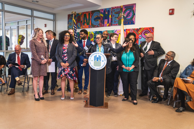 Gruppe von Menschen vor einem Rednerpult mit einem Mikrofon und einer Tafel mit der Aufschrift "Sancho-Stadtrat tritt dem Stadtrat bei", mit sitzenden und stehenden Teilnehmern, Flaggen, Feuerlöscher und Deckenleuchten im Hintergrund.