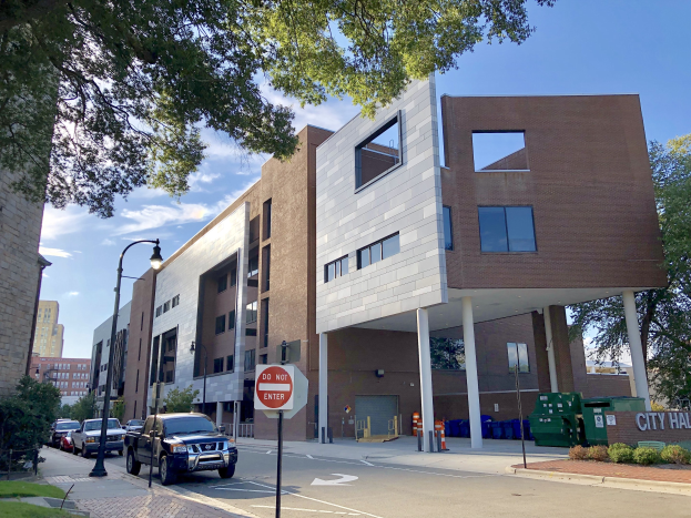 Großes mehrstöckiges Stadtzentrum Apartments Gebäude mit zahlreichen Fenstern, Straßeninfrastruktur einschließlich Pfosten, Lampen und Verkehrszeichen, umgeben von Fahrzeugen, Grünflächen und einem bewölkten Himmel.