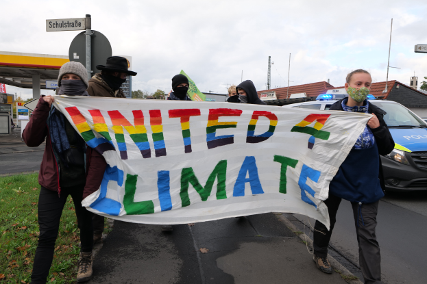 Eine Gruppe maskierter Personen mit einem "United Climate"-Schild vor einer Tankstelle, mit Fahrzeugen, Gras, Schildern, Gebäuden, Bäumen und einem bewölkten Himmel im Hintergrund.