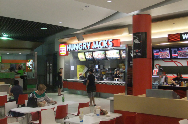 In einem Restaurant stehen Frauen an einer Essensbestellung auf einer Seite, während auf der anderen Seite Tische und Stühle angeordnet sind, an denen eine Frau und ein Mann an einem der Tische sitzen.