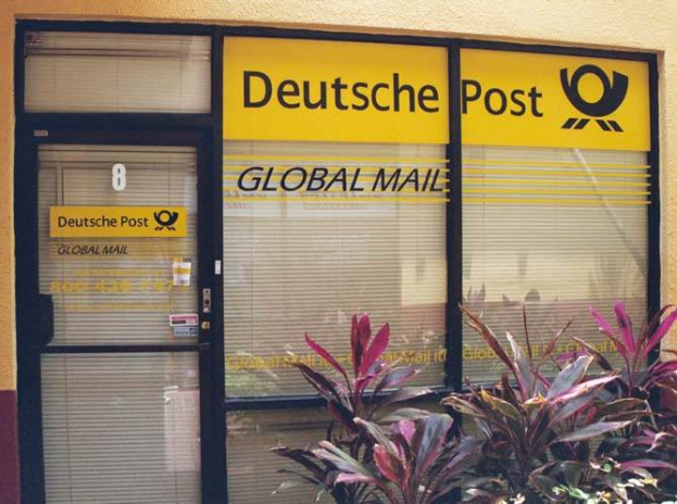 Ein Gebäude mit einer Tür, Jalousien und Pflanzen davor, mit einem Schild, auf dem "Deutsche Post Global Mail" steht.