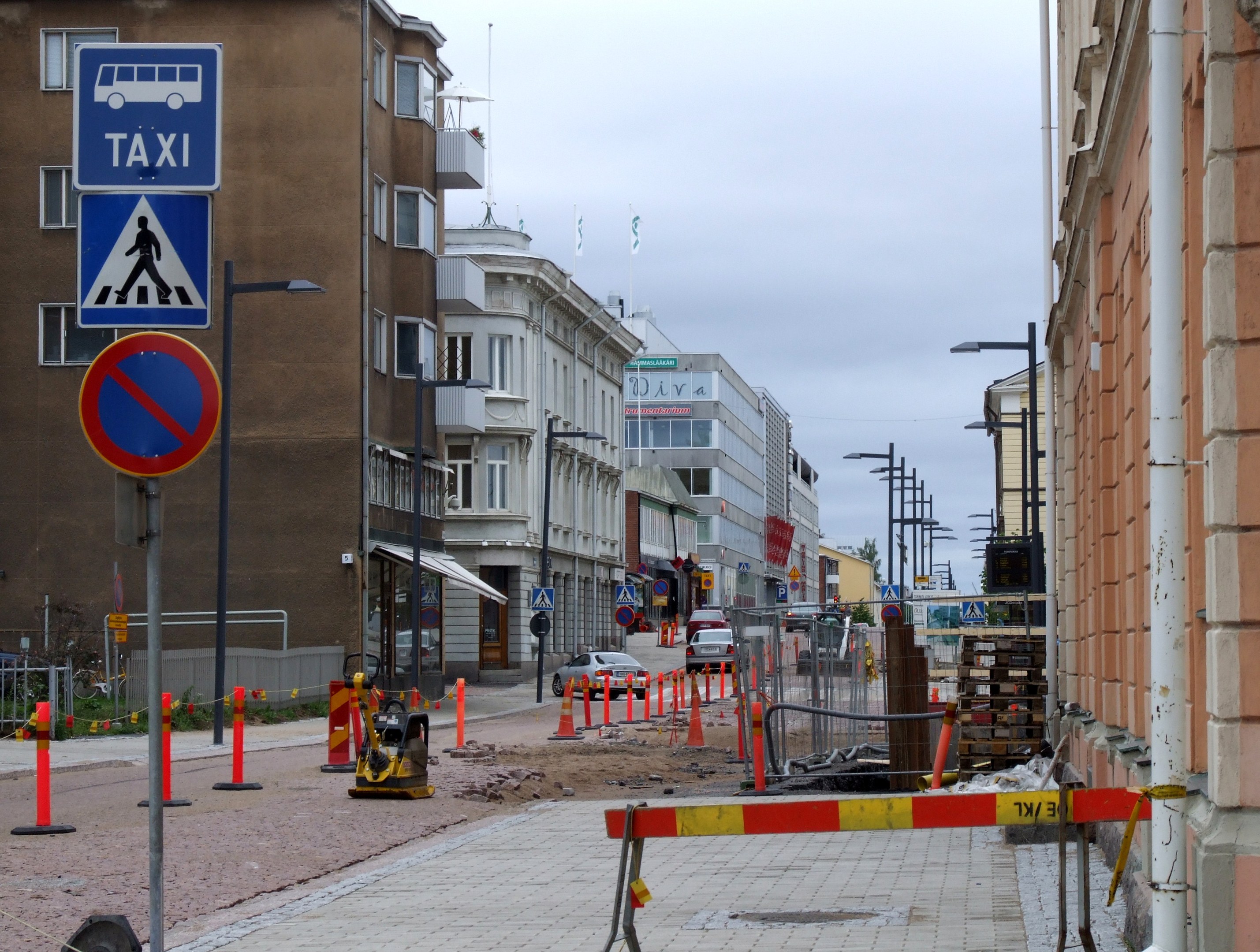 Eine Stadtstraße mit Gebäuden, Straßenlaternen, Verkehrsschildern, Verkehrsleitkegeln, Kraftfahrzeugen, Absperrpoller, Bäumen, einer Baustelle mit Verkehrsschildern und einem Himmel mit Wolken im Hintergrund.