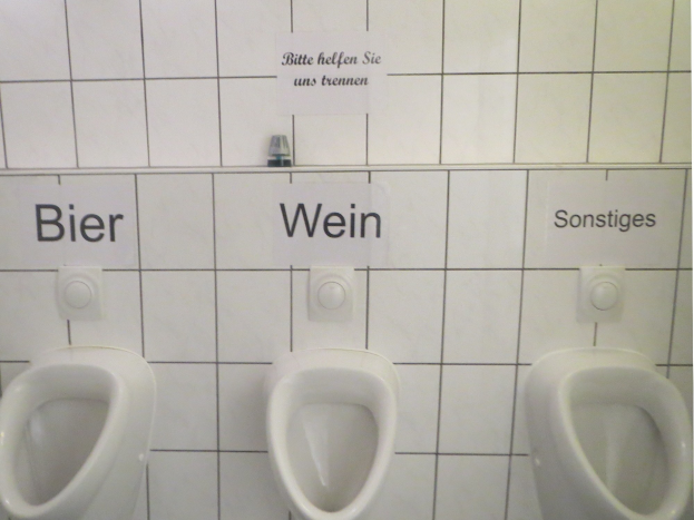 Drei weiße Toiletten an einer Wand montiert, jeweils mit einem Schild beschriftet, das 'Bier Wein' oder 'Sonstiges' liest.