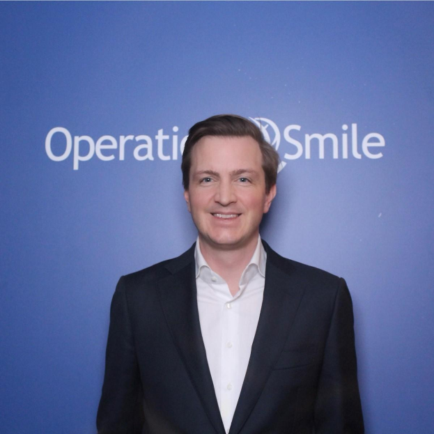 Ein Mann in einem Anzug lächelt vor einer blauen Wand mit "Operationsmile" darauf.