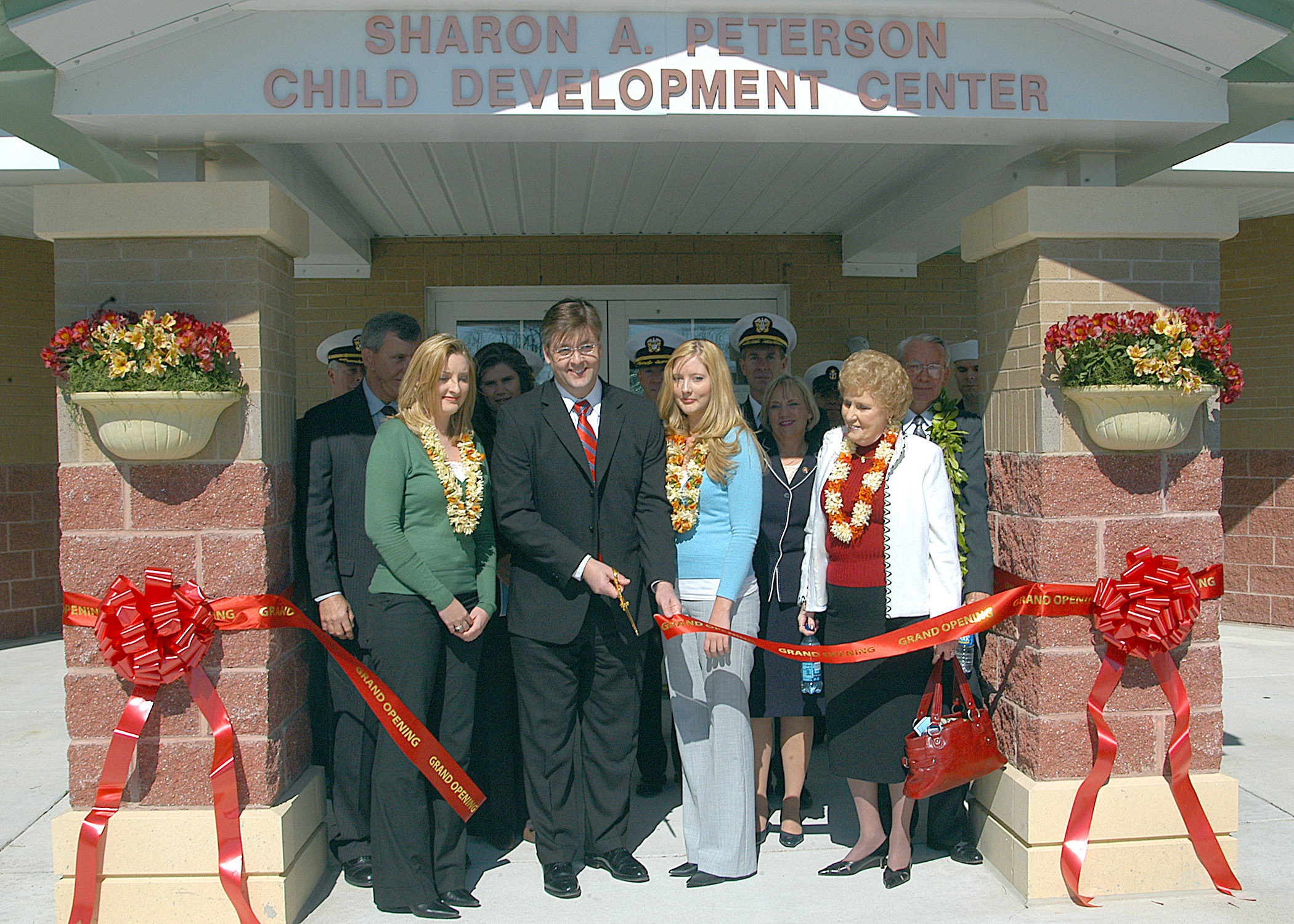 Eine Gruppe von Menschen steht vor dem Sharon A Peterson Child Development Center, einige tragen Kränze und eine Person hält eine Tasche, während Bänder an den Säulen und Blumentöpfe den Eingang säumen.