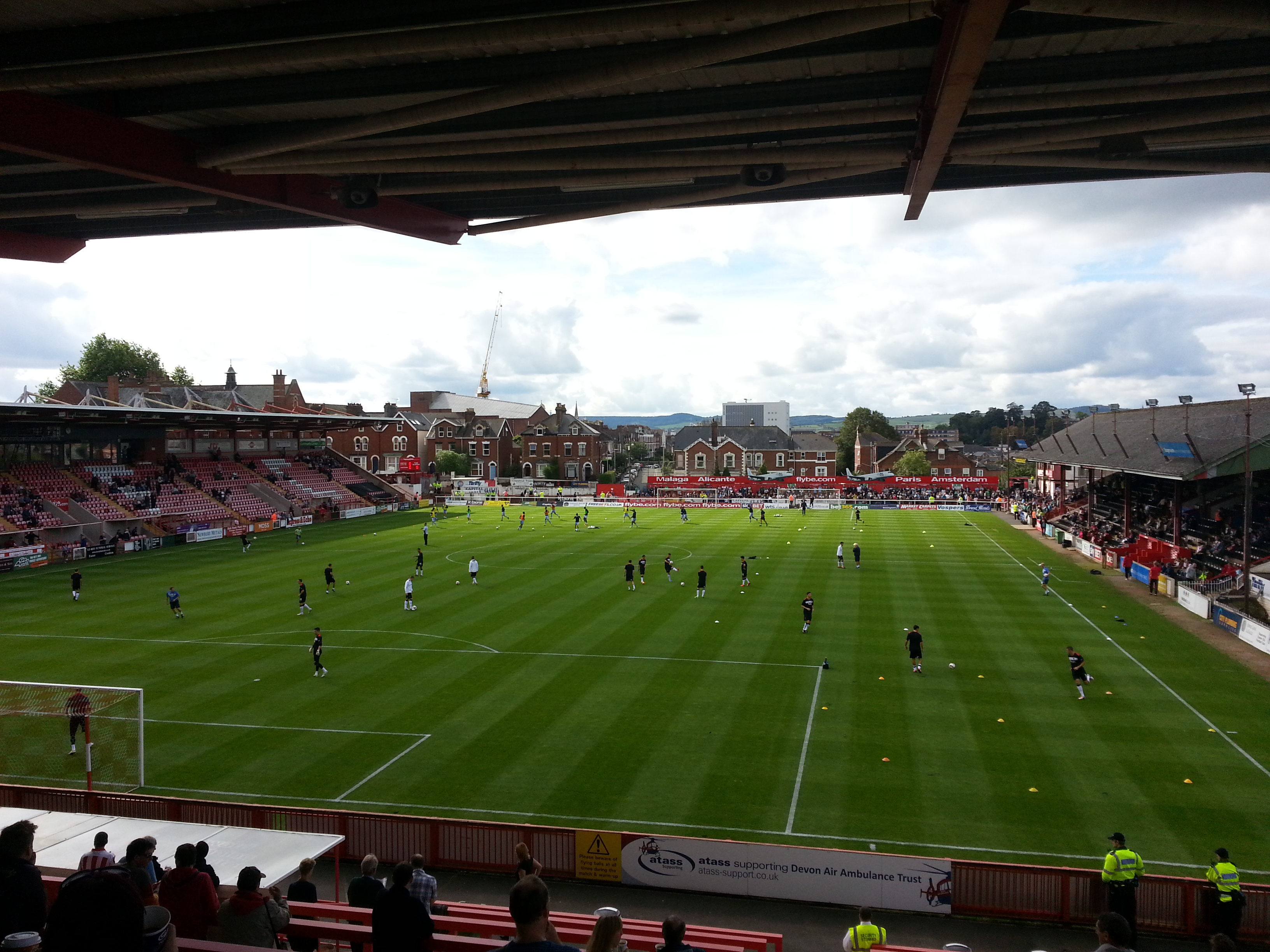 Ein Fußballspiel wird im St Mary's Stadium gespielt, mit Zuschauern auf den Bänken und Stehplätzen und einem Torpfosten auf der linken Seite sowie Gebäuden, Bäumen und einem bewölkten Himmel im Hintergrund.