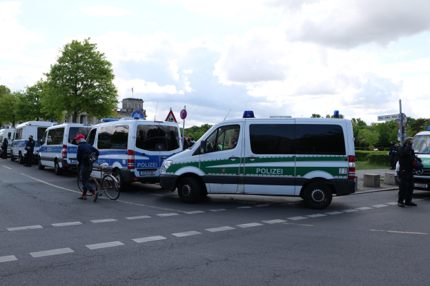 Eine Gruppe von Polizeiwagen, die entlang einer Straße geparkt sind, mit einem Radfahrer im Vordergrund und Gebäuden, Bäumen und Schildern im Hintergrund unter einem bewölkten Himmel.