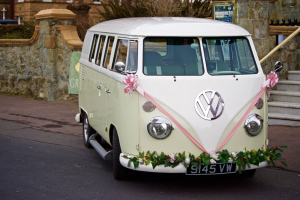 Ein weißer und pinker VW Camper mit floralen Verzierungen, der vor einer Steinmauer, einer Treppe und einem Gebäude geparkt ist.