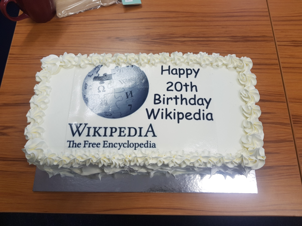 Geburtstagstorte mit "Happy 20th Birthday Wikipedia" darauf, auf einem Tisch mit einer Tasse, einer Hülle und anderen Gegenständen.