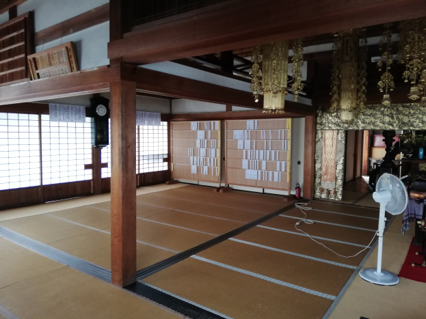 Innenraum eines traditionellen japanischen Hauses mit Tatami-Matten, Tischventilator, Teppich, Feuerlöscher, Gegenständen auf Oberflächen, Uhr, Papieren an der Wand, Holzpfosten, Dach mit Deckenleuchten und einem Kronleuchter.