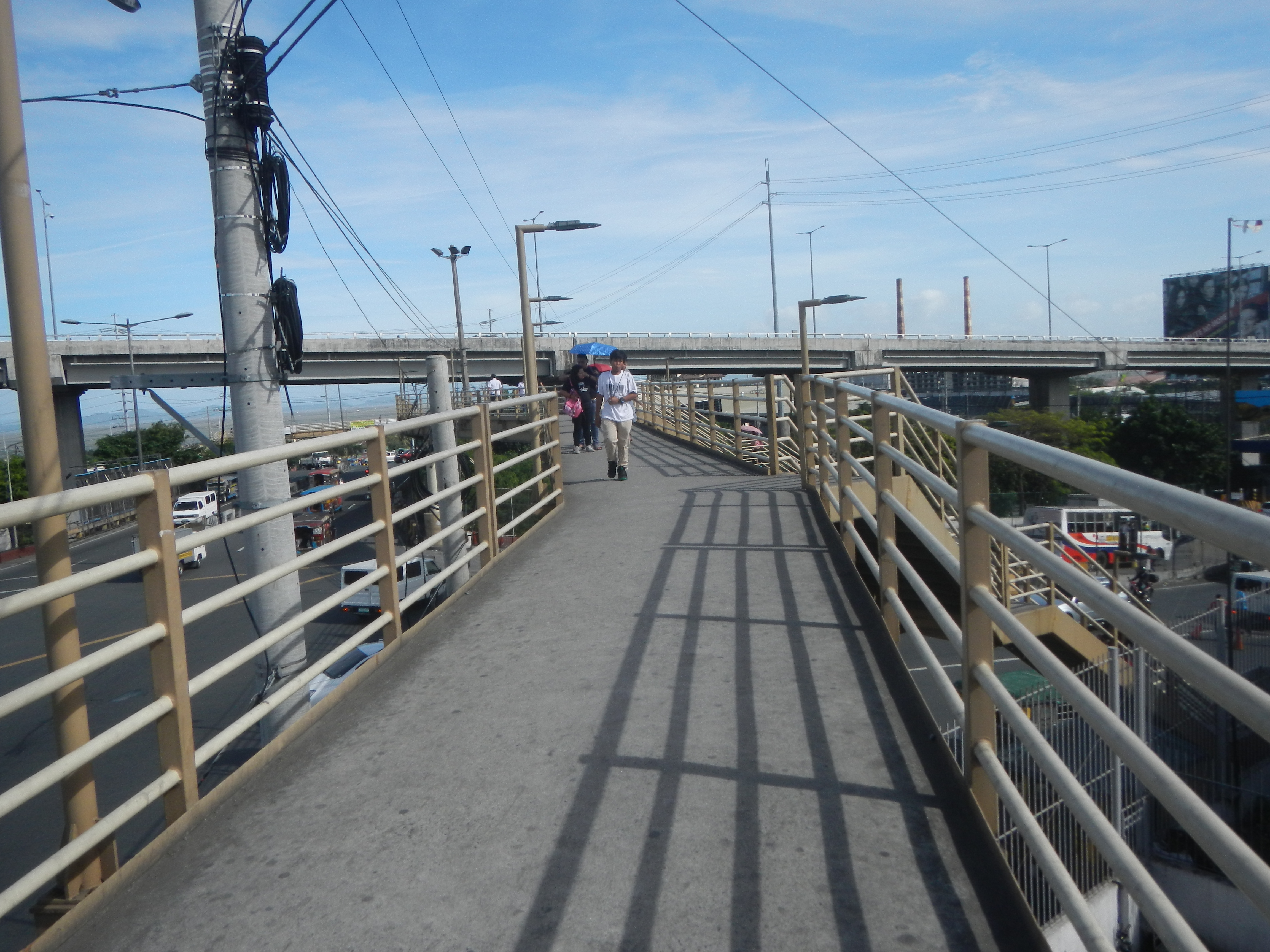 Eine Fußgängerüberquerung mit Menschen, die überqueren, umgeben von Geländern, Laternen, Strommästen mit Drähten, Fahrzeugen auf der Straße, Bäumen, Gebäuden und einem bewölktem Himmel im Hintergrund.