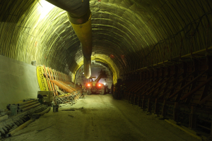Großer Tunnel im Bau an einer Baustelle mit Fahrzeugen, verstreuten Materialien, einer Wand auf der linken Seite, Überkopfrohren und Hintergrundbeleuchtung.