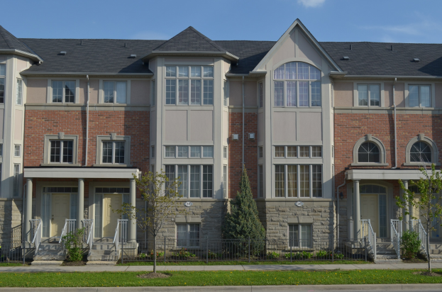 Reihe von Stadthäusern mit Fenstern, Türen, Säulen, Geländern, Treppen, Pflanzen, Bäumen, Gras und einer Straße im Vordergrund, unter einem Himmel-Hintergrund.