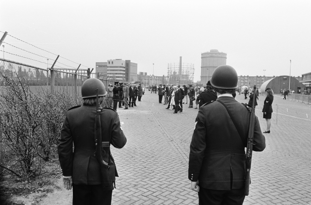 Zwei Polizisten in Helmen und mit Waffen in den Händen stehen nebeneinander auf einem Bürgersteig, links daneben ein Zaun und Pflanzen, im Hintergrund Gebäude, Pfähle und einen klaren Himmel.