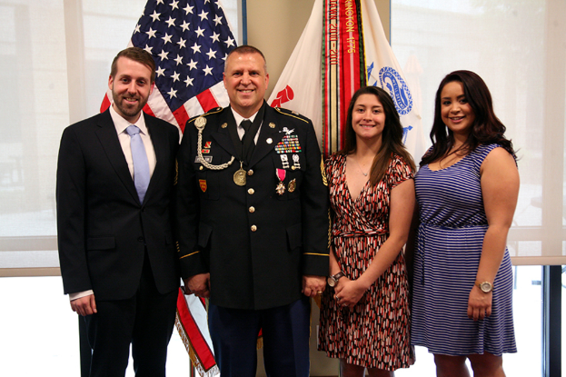 Zwei Frauen in Kleidern, eine Person in Uniform und ein Mann in einem Anzug, der drinnen steht und lächelt, mit Jalousien und Flaggen im Hintergrund.