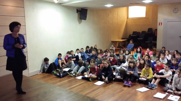 Eine Gruppe von Kindern, Jungen und Mädchen, sitzt auf dem Boden mit Büchern vor sich, während eine Frau auf der linken Seite auf der Bühne steht.