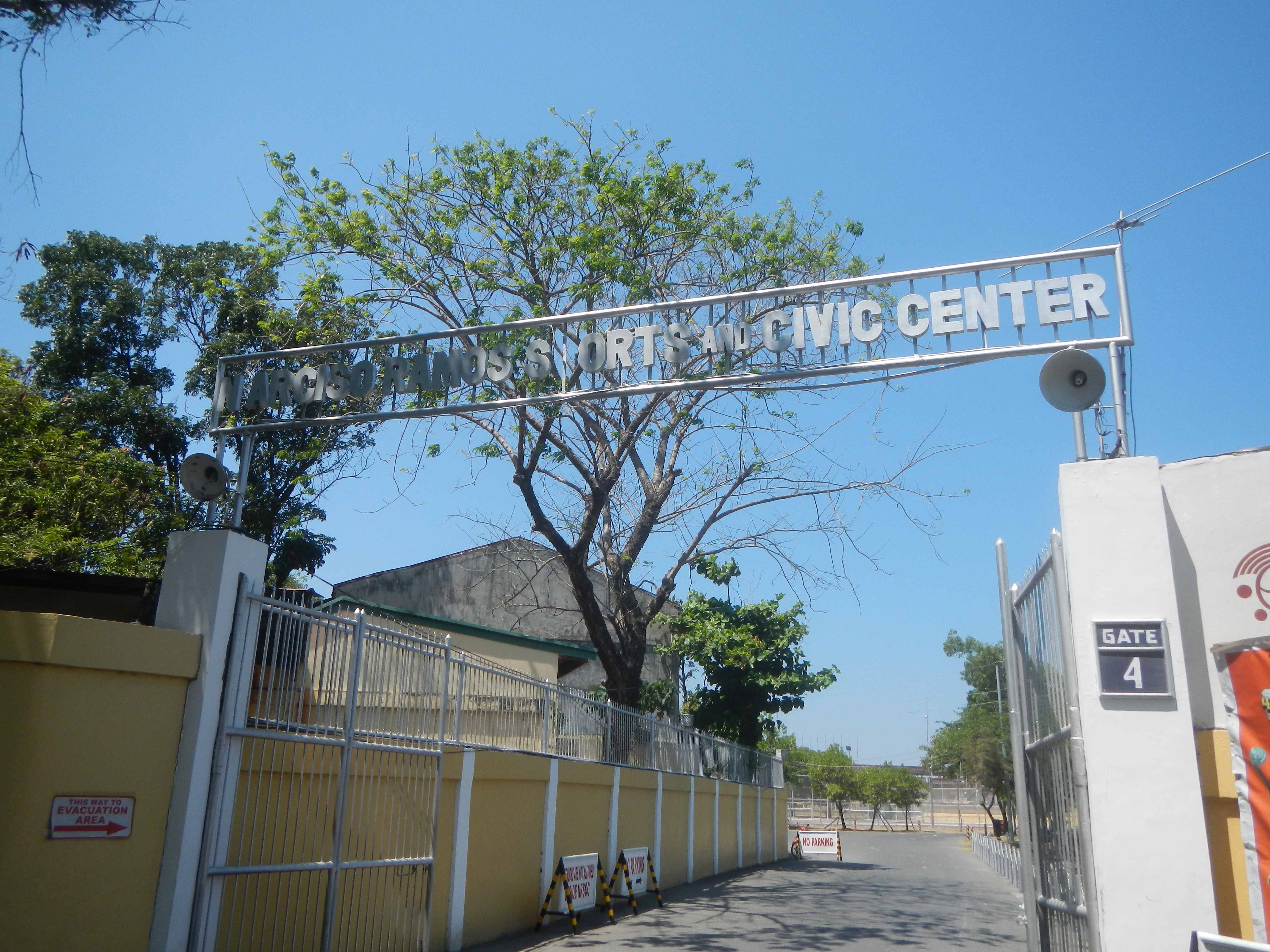 Der Eingang zu einem Sport- und Bürgerzentrum zeigt einen Bogen mit Text, eine Wand mit einem Tor, eine Fahne mit Text und Zahlen auf der rechten Seite, eine Straße unten, Bäume und Gebäude im Hintergrund und einen klaren blauen Himmel darüber.