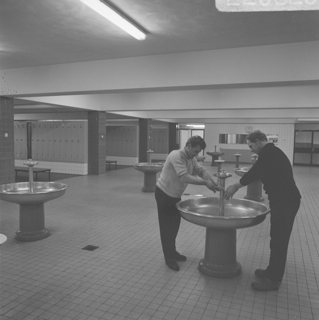 Zwei Männer, die nebeneinander in einer öffentlichen Toilette mit Waschbecken, Säulen, Wänden, Türen und Lampen stehen, in Schwarz-Weiß dargestellt.